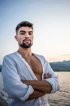 Strong attractive muscular caucasian bearded man looks serious at the camera with white shirt open and strong chest with the pectoral - Portrait of male sexy young model with deep and intense gaze