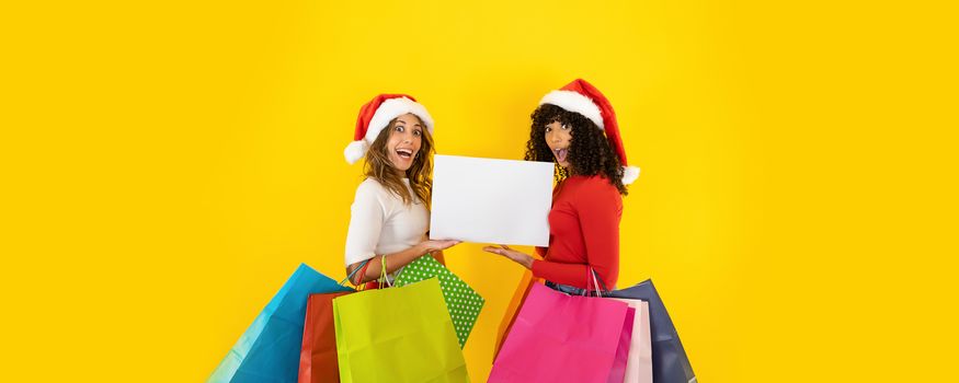 Two young cute women wearing Santa Claus hat looking to the camera with astonished face, open mouth, wide eyes, holding colored shopping bags - Christmas banner with yellow background for copy space