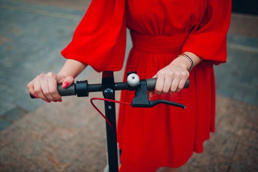 Young woman holds steering wheel of electric scooter in red dress at the city.