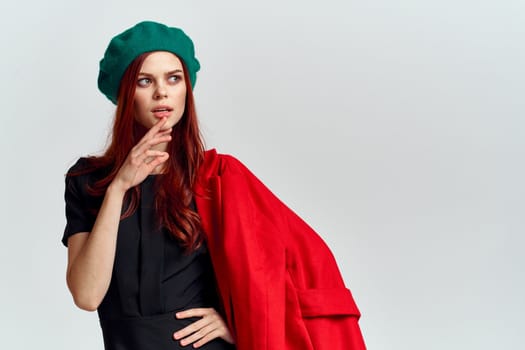 A woman holds in her hand a red coat and a dark T-shirt Green hat cropped view of a light background. High quality photo