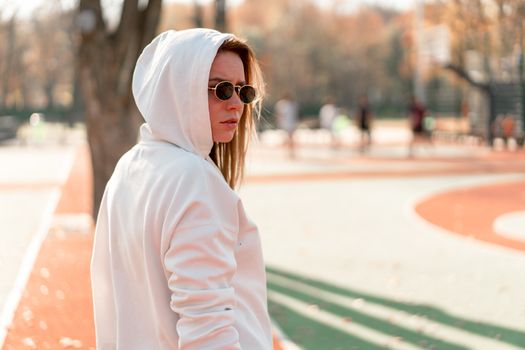 Outdoor close up portrait of young beautiful woman with in glasses, dressed in a white sweater, near the sportsground. Woman in a hood turned her head to the camera. White hoodie