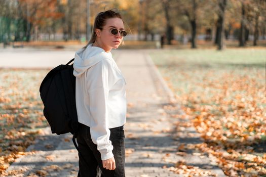 A young beautiful woman with a ponytail and sunglasses, with a backpack on his shoulders in the park. Photo from the back. White hoodie