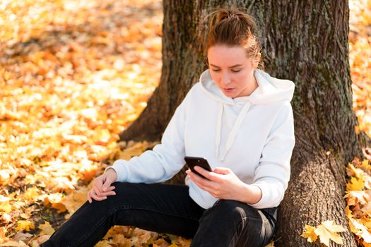 Woman in a white hoodie sweater with a hood sits on ground in the park and holding a cellphone in her hands. cute girl use smartphone for communication Female texting a message Communicaion concept