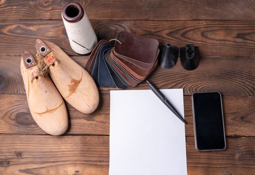 Leather samples for shoes and wooden shoe last on dark wooden table with Empty white sheet of paper for notes.. Designer furniture clothes. Shoe maker workspace.