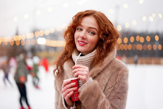 Young beautiful redhead girl freckles ice rink background Pretty woman curly hair portrait walking new year fair Modern cute female drink hot beverage mulled wine chrismas holiday decorated street.