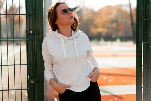 Outdoor close up portrait of young beautiful woman with long hair in sunglasses, dressed in a white hoodie sweater, near the sportsground. youth culture summer pastime