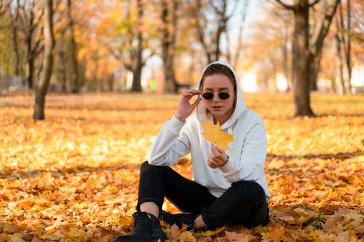 Woman in a white sweater with a hood sits on ground in the park and holding a leaf in her hands. Cute girl in white hoodie is sitting on the yellow leaves carpet