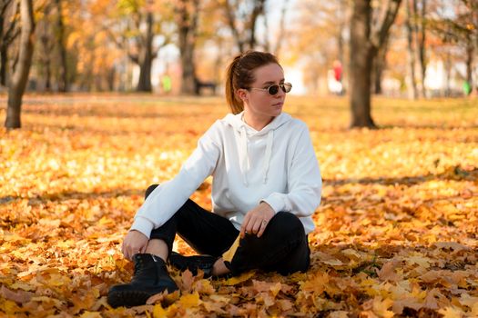 Woman in a white sweater with a hood sits on ground in the park and holding a leaf in her hands. Cute girl in white hoodie is sitting on the yellow leaves carpet