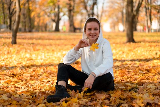 Woman in a white sweater with a hood sits on ground in the park and holding a leaf in her hands. Cute girl in white hoodie is sitting on the yellow leaves carpet
