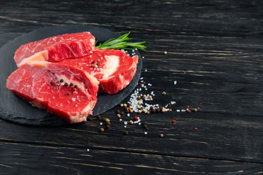 Three pieces of juicy raw beef with rosemary on a stone cutting board on a black wooden table background. Meat for barbecue or grill sprinkled with pepper and salt seasoning