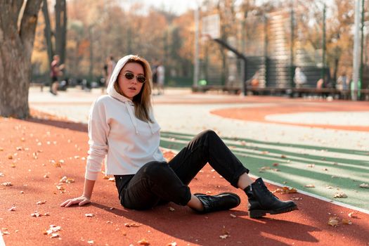 Outdoor portrait of young beautiful woman with long in sunglasses and a white hooded sweater sitting on the sportsground track. youth culture summer pastime