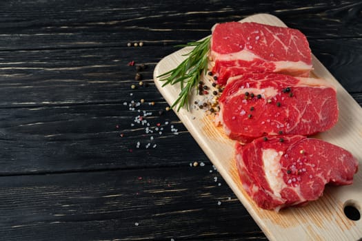 Three pieces of juicy raw beef with rosemary on a cutting board on a black wooden table background. Meat for barbecue or grill sprinkled with pepper and salt seasoning