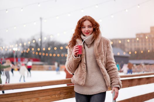 Young beautiful redhead girl freckles ice rink background Pretty woman curly hair portrait walking new year fair Modern cute female skate ice rink chrismas holiday decorated street.