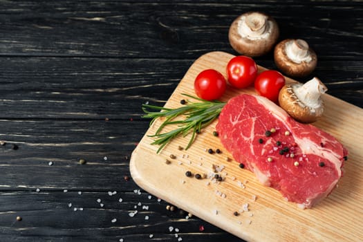 One pieces of juicy raw beef with rosemary on a cutting board on a black wooden table background. Meat for barbecue or grill next to cherry tomatoes sprinkled with pepper and salt seasoning