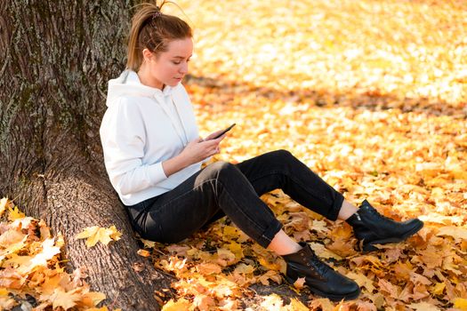 Woman in a white hoodie sweater with a hood sits on ground in the park and holding a cellphone in her hands. cute girl use smartphone for communication Female texting a message Communicaion concept