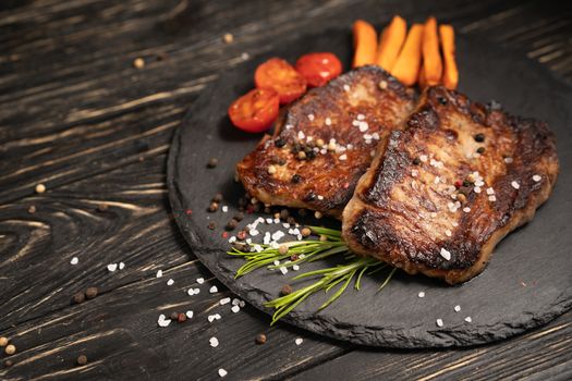 A juicy piece of fried meat with grilled cherry tomatoes and carrot lies on a stone plate against a black wooden table. Degree of roasting well done. Food concept with copy space