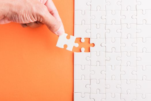 Top view flat lay of hand-holding last piece white paper jigsaw puzzle game last pieces put to place for solve problem complete mission, studio shot on an orange background, quiz calculation concept