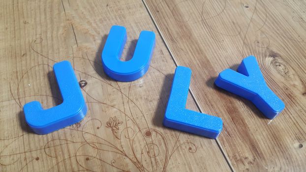 Plastic colored alphabets making words July are placed on a wooden floor. These plastic letters can be used for teaching kids.