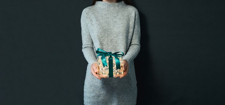 Woman in gray pullover holding luminous gift box in hands on dark background. Glowing yellow light bulbs into transparent gift box with dark green ribbon. Christmas and New Year concept