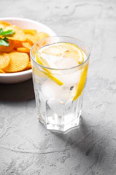 Fresh ice cold water drink with lemon near to fried corrugated golden potato chips with parsley leaf in wooden bowl on concrete background, angle view