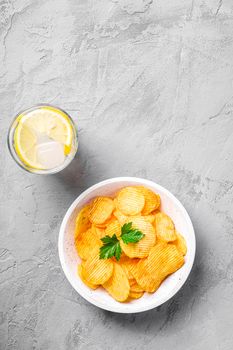 Fresh ice cold water drink with lemon near to golden potato chips with parsley leaf in wooden bowl on concrete background, top view copy space