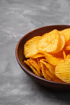 Fried corrugated golden potato chips in brown wooden bowl on concrete background, angle view