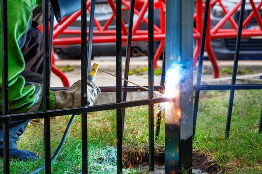 Hands of a welder in protective gloves with an electrode weld a metal fence at a construction site, bright sparks, blue smoke fly, selective focus, copy space.