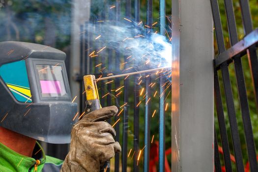 A welder in a protective helmet and gloves using an electrode welds a metal fence in a park area, bright sparks, blue smoke fly, selective focus, copy space.