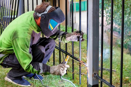 A welder wearing protective clothing, helmet and gloves using an electrode welds a metal fence around the work area, setting up a fence, selective focus, copy space.