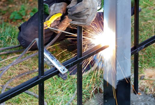 Bright sparks from electric welding fly in different directions against the background in the blurring of the working welder and grass.