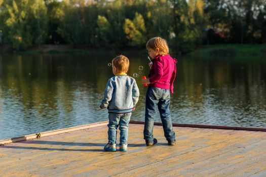 A little boy and a girl blow bubbles and stand on a wooden flooring near a pond in a city park one autumn evening