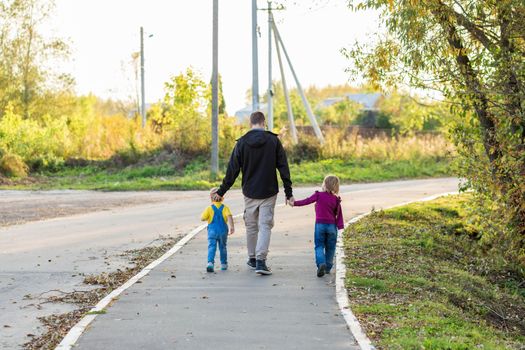 Dad leads the hands of the children along the sidewalk in the village one autumn evening