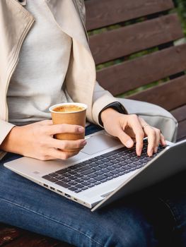 Business woman sits on wooden bench in urban park with laptop and cardboard cup of coffee. Student learns remotely from outdoors. Modern Internet technologies.