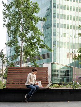 Business woman with short haircut and eyeglasses sits on bench in urban park with laptop and cardboard cup of coffee. Student learns remotely from outdoors. Modern Internet technologies. Video call.