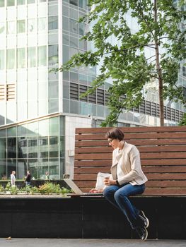 Business woman with short haircut and eyeglasses sits on bench in urban park with laptop and cardboard cup of coffee. Student learns remotely from outdoors. Modern Internet technologies. Video call.