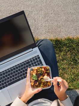 Woman sits on park bench with laptop and take away lunch box. Healthy bowl with vegetables. Casual clothes, urban lifestyle of millennials. Healthy nutrition.