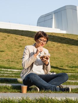 Woman sits on park bench with take away lunch box, cardboard cup of coffee. Healthy bowl with vegetables. Casual clothes, urban lifestyle of millennials. Healthy nutrition.