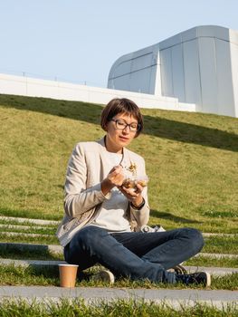 Woman sits on park bench with take away lunch box, cardboard cup of coffee. Healthy bowl with vegetables. Casual clothes, urban lifestyle of millennials. Healthy nutrition.