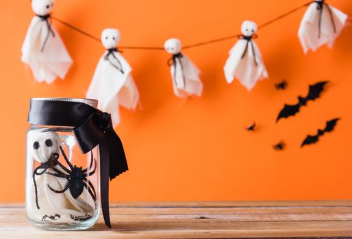 Funny Halloween day decoration party, Baby white ghost crafts scary face in jar glass on wood table, studio shot isolated on orange background have spider and bats, Happy holiday DIY concept