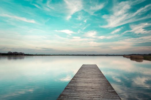 A wooden jetty towards calm water and clouds against a blue sky