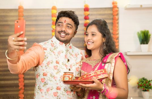 Indian couple in traditional dress taking selfies after celebrating Karva Chauth festival at home - Concept of using Mobile, technology during festival celebrations