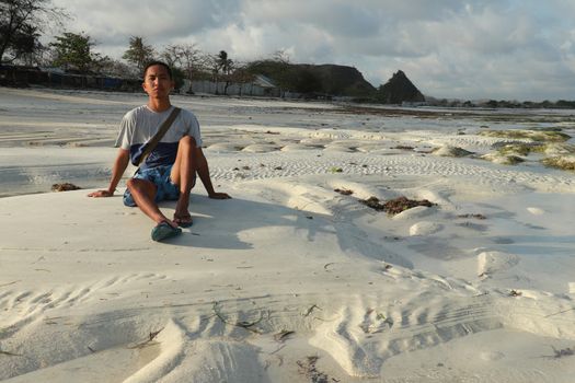 Teenager sitting on white sand at Tanjung Aan beach, Lombok, Indonesia. A young Asian man looks at the landscape.