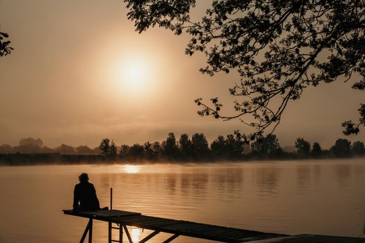 silhouette woman at wooden jetty during sunset in the early morning over the river maas in limburg in holland with the trees mist and hazy fog