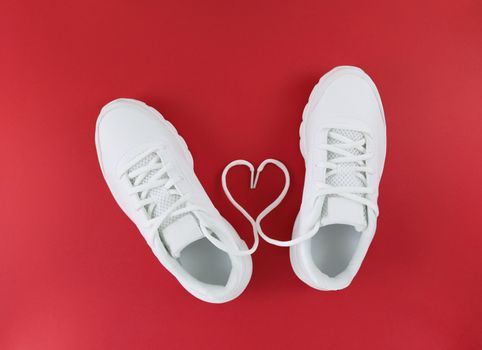 White sports shoes and heart shape from laces on red background. Simple flat lay.