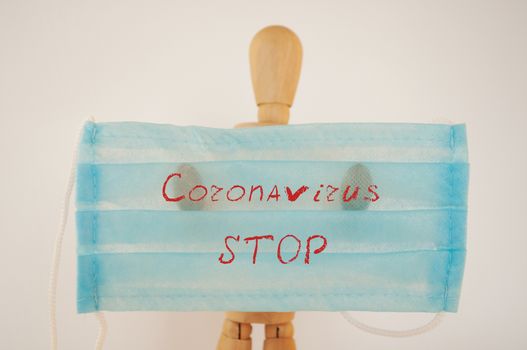 Surprised wooden man holds a blue medical protective mask with the inscription: stop, coronavirus. The concept of protection during the coronavirus pandemic. Copy space