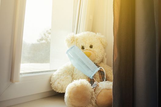 Coronavirus epidemic, COVID-19. The bear sits in a medical mask on the windowsill and looks at the camera. The concept of worldwide isolation is driven by a pandemic. Stay at home.