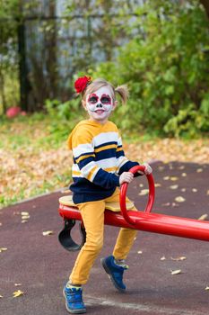 A little preschool girl with Painted Face, rides on a swing in the playground, celebrates Halloween or Mexican Day of the Dead.