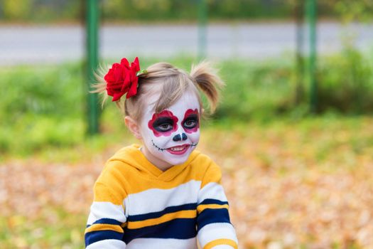 A little preschool girl with Painted Face, smiling at the camera on the playground, celebrates Halloween or Mexican Day of the Dead..