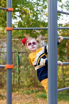 A little preschool girl with Painted Face, climbed the Swedish wall in the playground, celebrates Halloween or Mexican Day of the Dead.