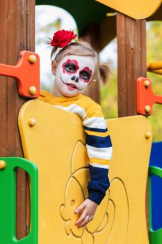 A little preschool girl with Painted Face, climbed the slide on the playground, celebrates Halloween or Mexican Day of the Dead.